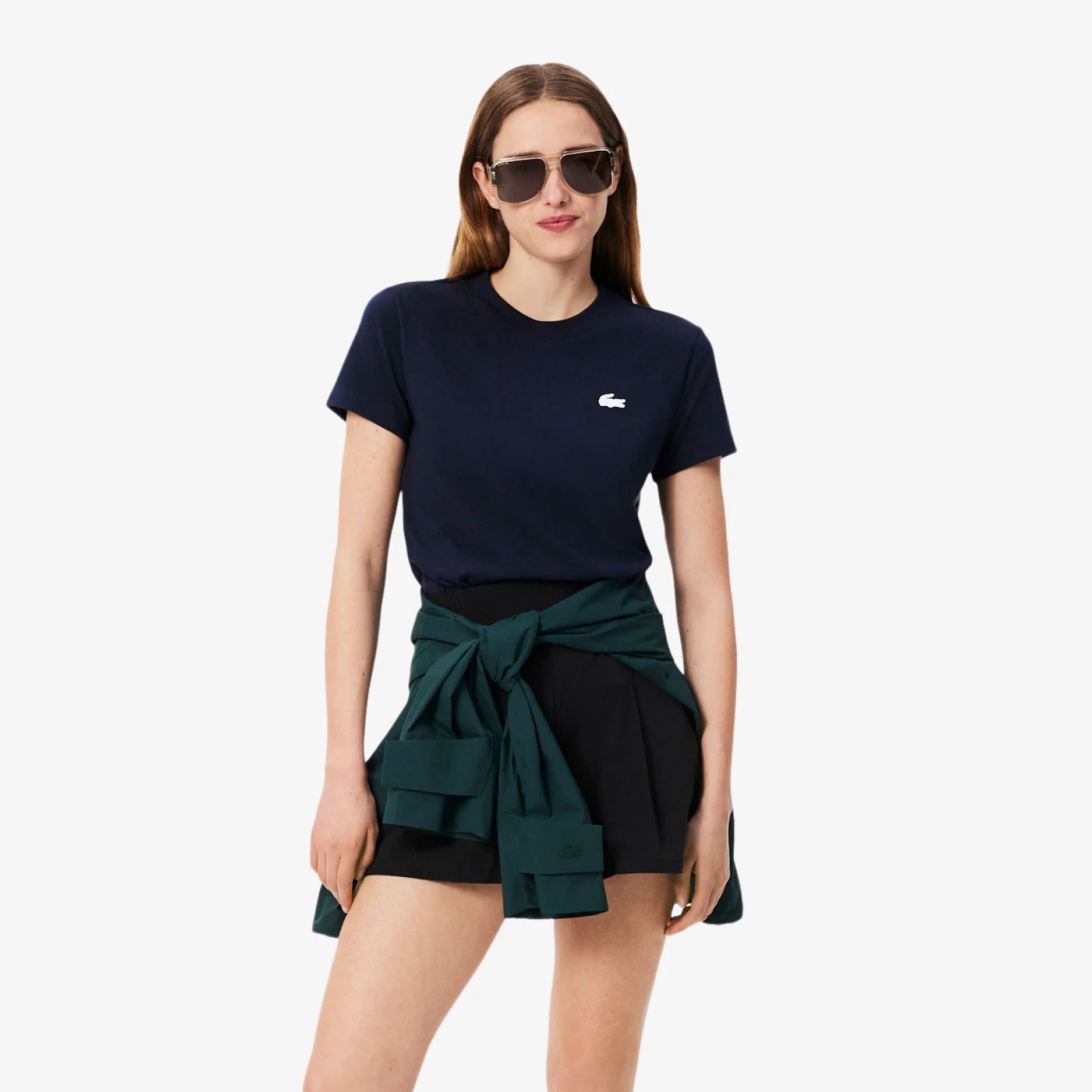 model is wearing sunglasses, a white Lacoste hoodie, a dark green Lacoste rain jacket, black Lacoste skort and white/pink Lacoste L003 sneakers